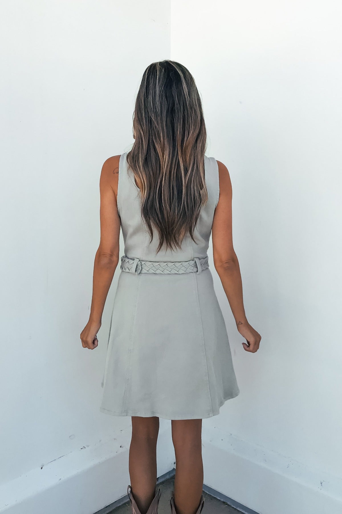 A woman with long, wavy hair faces a white corner in the City Slate Belted Mini Dress and boots.