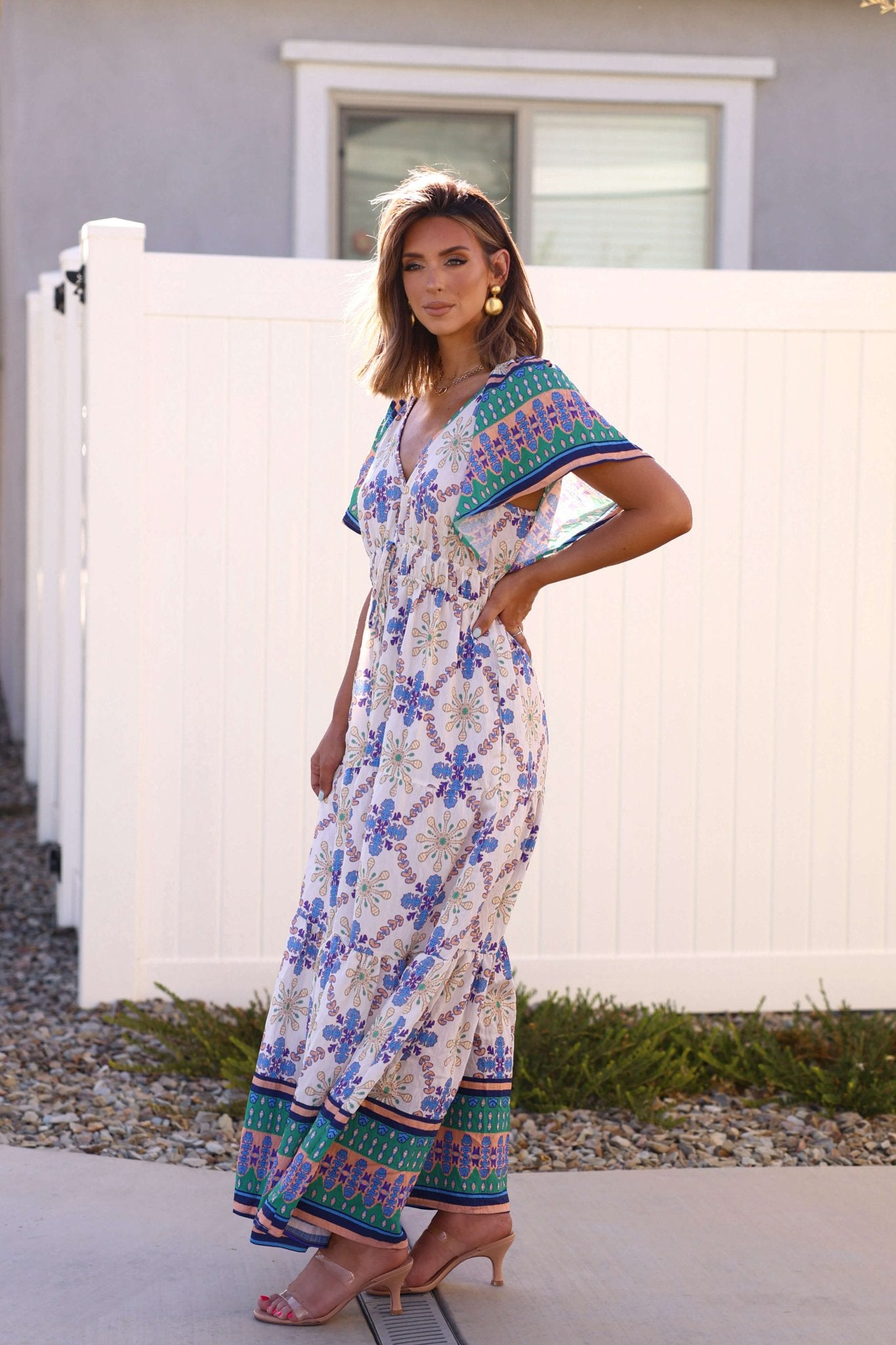 Woman stands outdoors by a white fence, wearing the Claire Blue Print Tiered Maxi Dress - DOORBUSTER and nude heels.