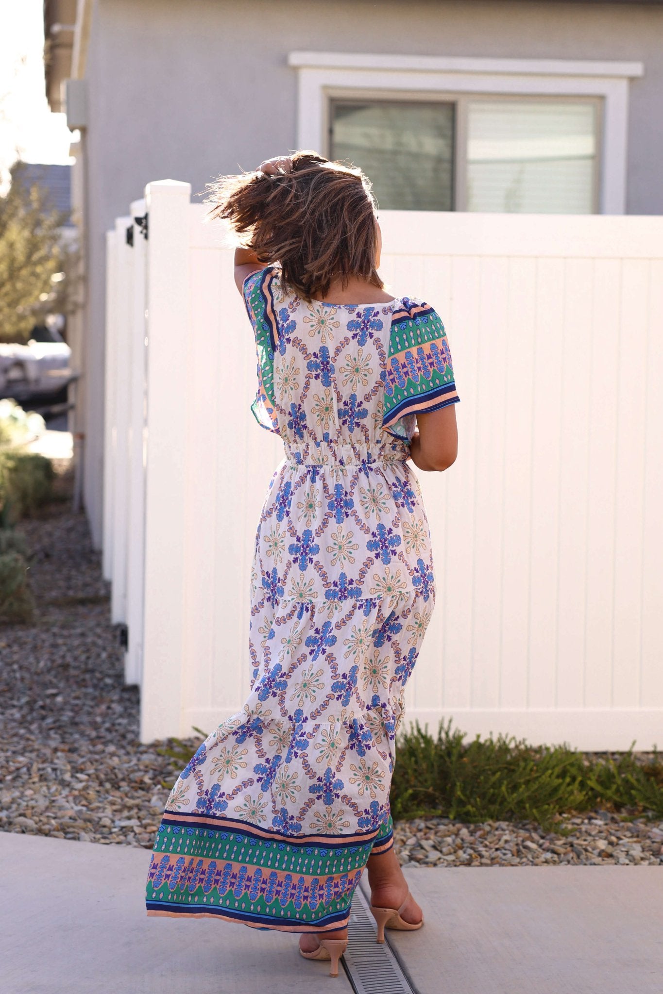 A woman in the Claire Blue Print Tiered Maxi Dress - DOORBUSTER walks outside on a sunny day, her hair blowing in the wind.
