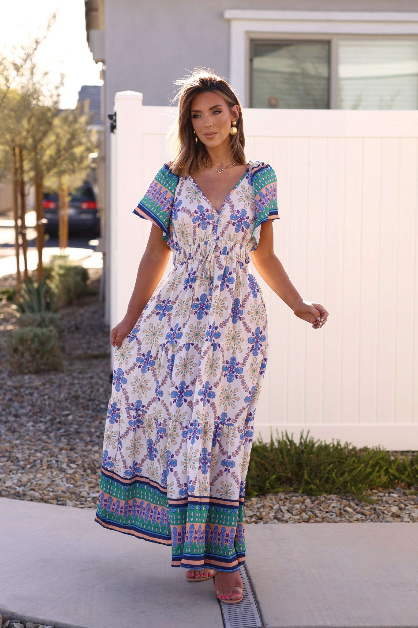 Woman wearing the Claire Blue Print Tiered Maxi Dress - DOORBUSTER, standing outdoors by a white fence and lush plants.