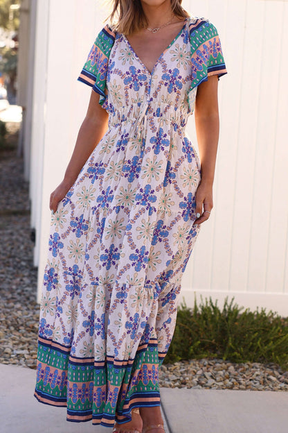 Woman in the Claire Blue Print Tiered Maxi Dress - DOORBUSTER, a flowy, short-sleeve V-neck dress, stands outdoors on a sunny day.
