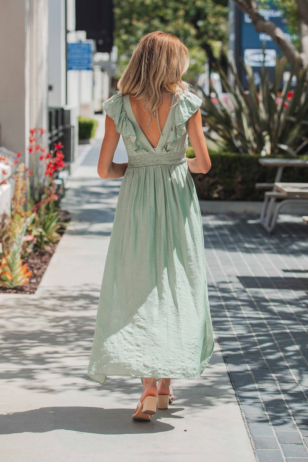 A woman in the Classic Sage Ruffled Midi Dress with a smocked waist walks outside on a sunny day, her back to the camera.