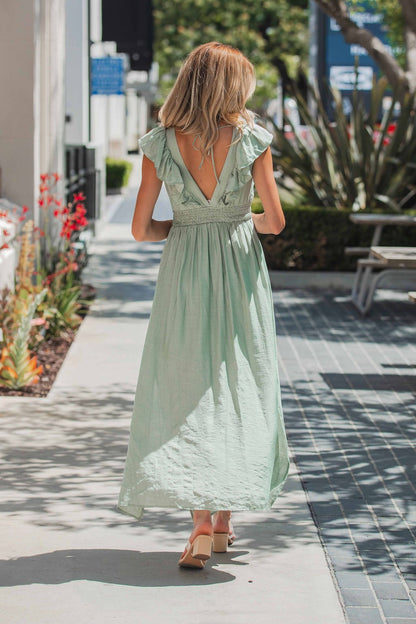 A woman in the Classic Sage Ruffled Midi Dress with a smocked waist walks outside on a sunny day, her back to the camera.