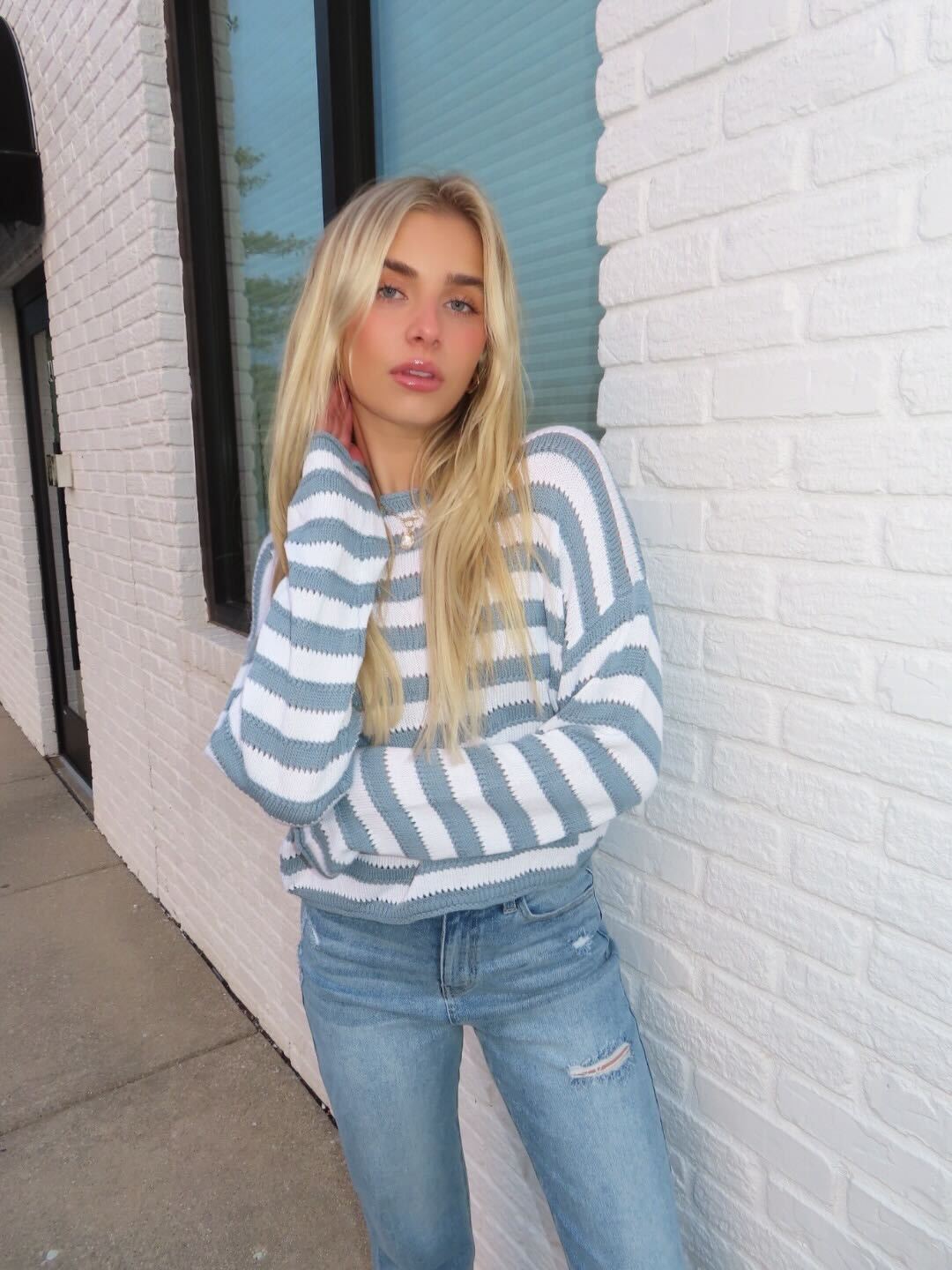 A young woman stands by a white brick wall, wearing a Cloud Grey and White Striped Sweater and light blue jeans.