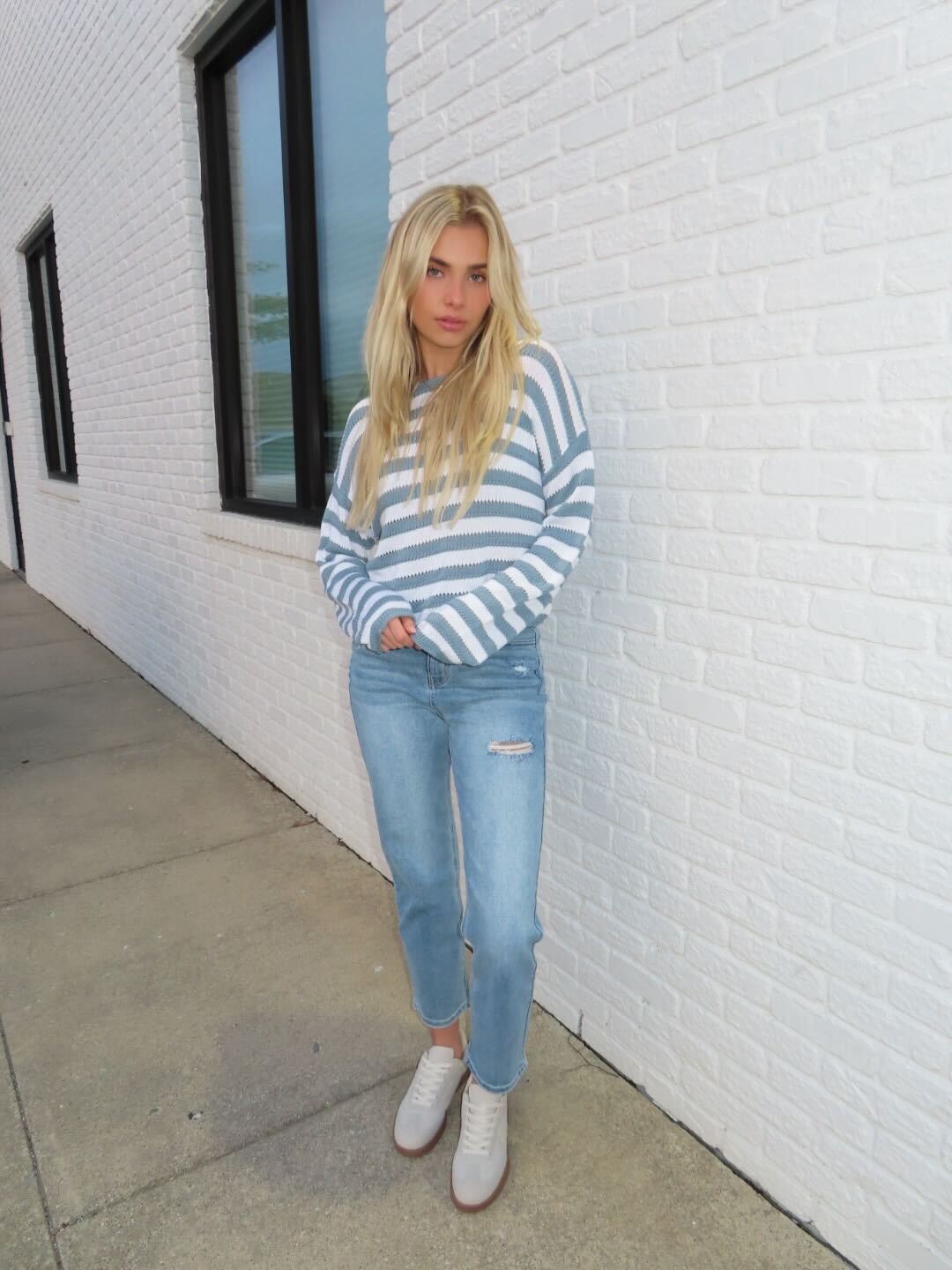 A woman wears a Cloud Grey and White Striped Sweater with jeans, standing by a white brick wall for a classic sidewalk style.