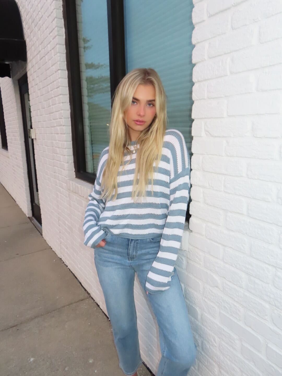 A young woman in a Cloud Grey and White Striped Sweater and jeans stands by a white brick wall, showcasing a classic style.