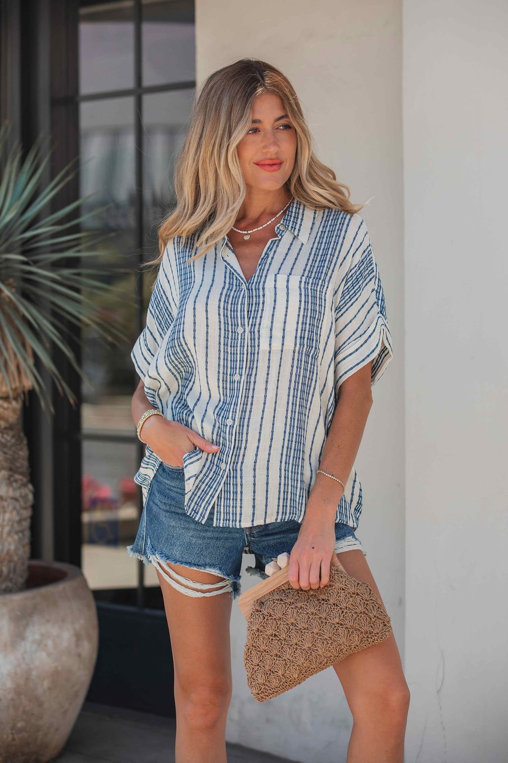 Woman in a Coastal Ease Striped Top - Navy, with a collared V neckline and denim shorts, holding a woven clutch by a potted plant.