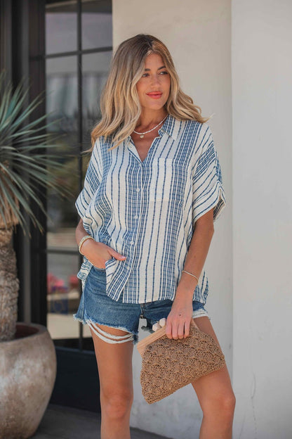 Woman in a Coastal Ease Striped Top - Navy, with a collared V neckline and denim shorts, holding a woven clutch by a potted plant.