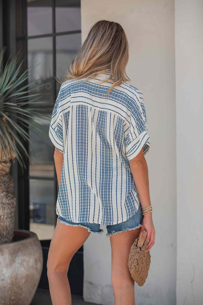 A woman stands outside, facing away, wearing the Coastal Ease Striped Top - Navy with denim shorts and holding a textured clutch.