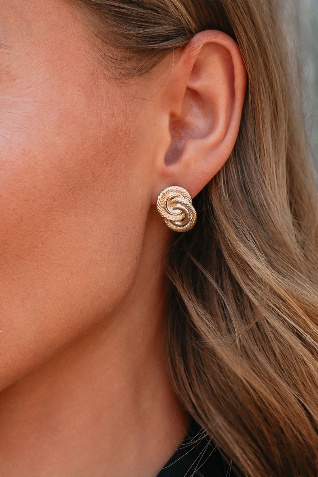 Close-up of a woman's ear wearing Coastal Gold Interlink Earrings; her wavy brown hair is visible.