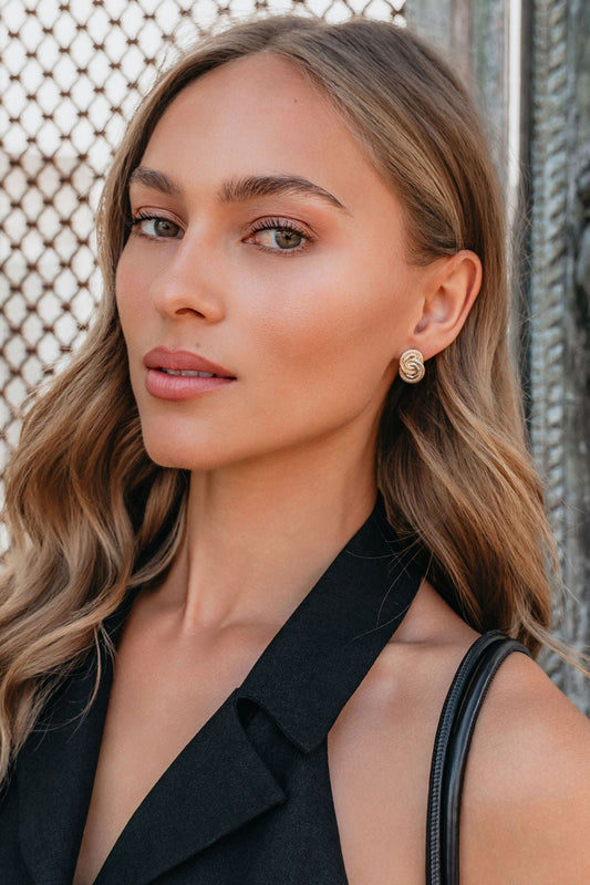 Woman with wavy blonde hair wearing a black sleeveless top and Coastal Gold Interlink Earrings, in front of a patterned metal background.