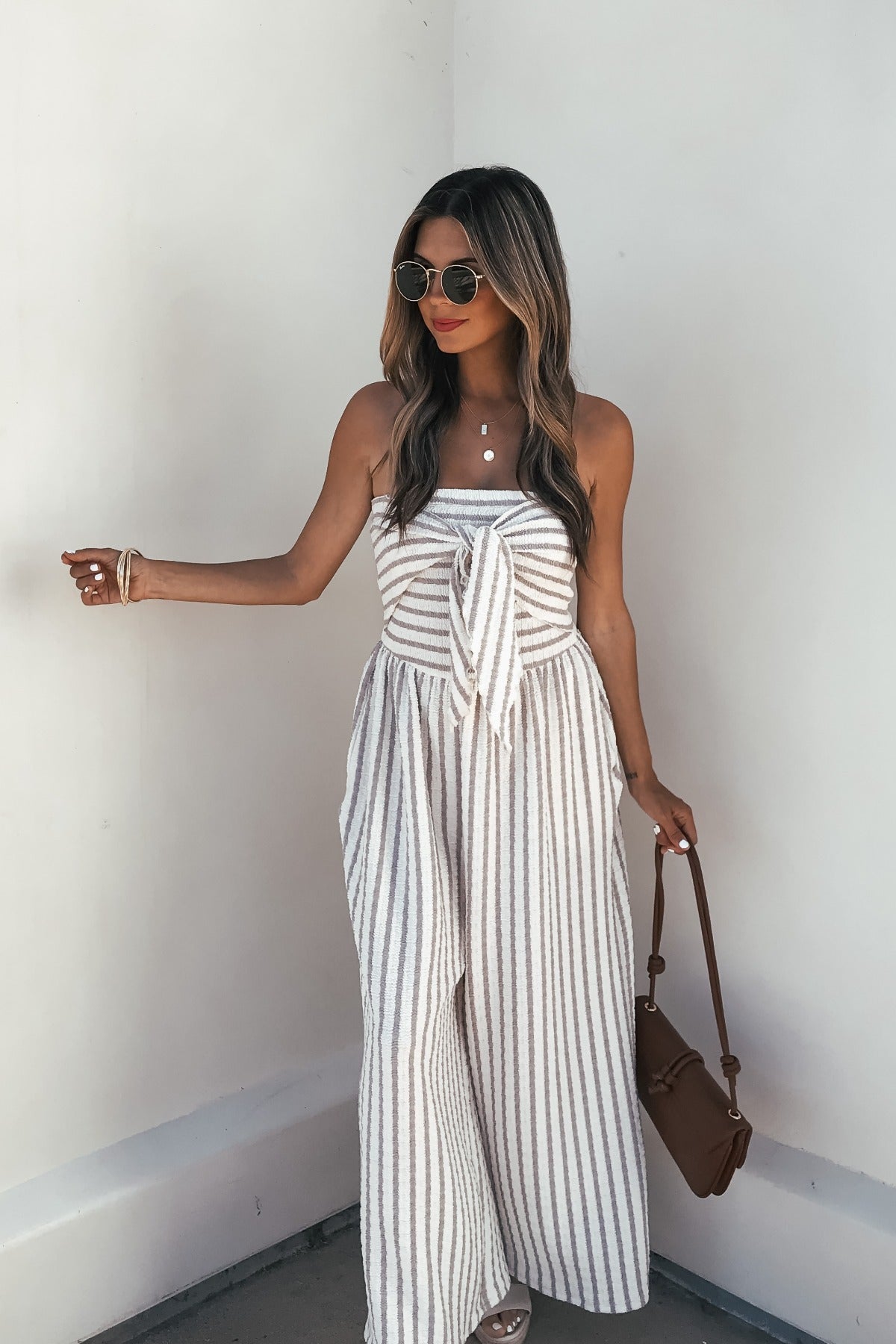 Woman in the Coastal Striped Wide Leg Jumpsuit - Taupe outfits with sunglasses and a brown bag, standing by a white wall.