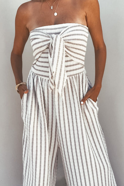 Woman wears the Coastal Striped Wide Leg Jumpsuit – Taupe with a strapless neckline, front tie, pockets, and breezy wide legs.