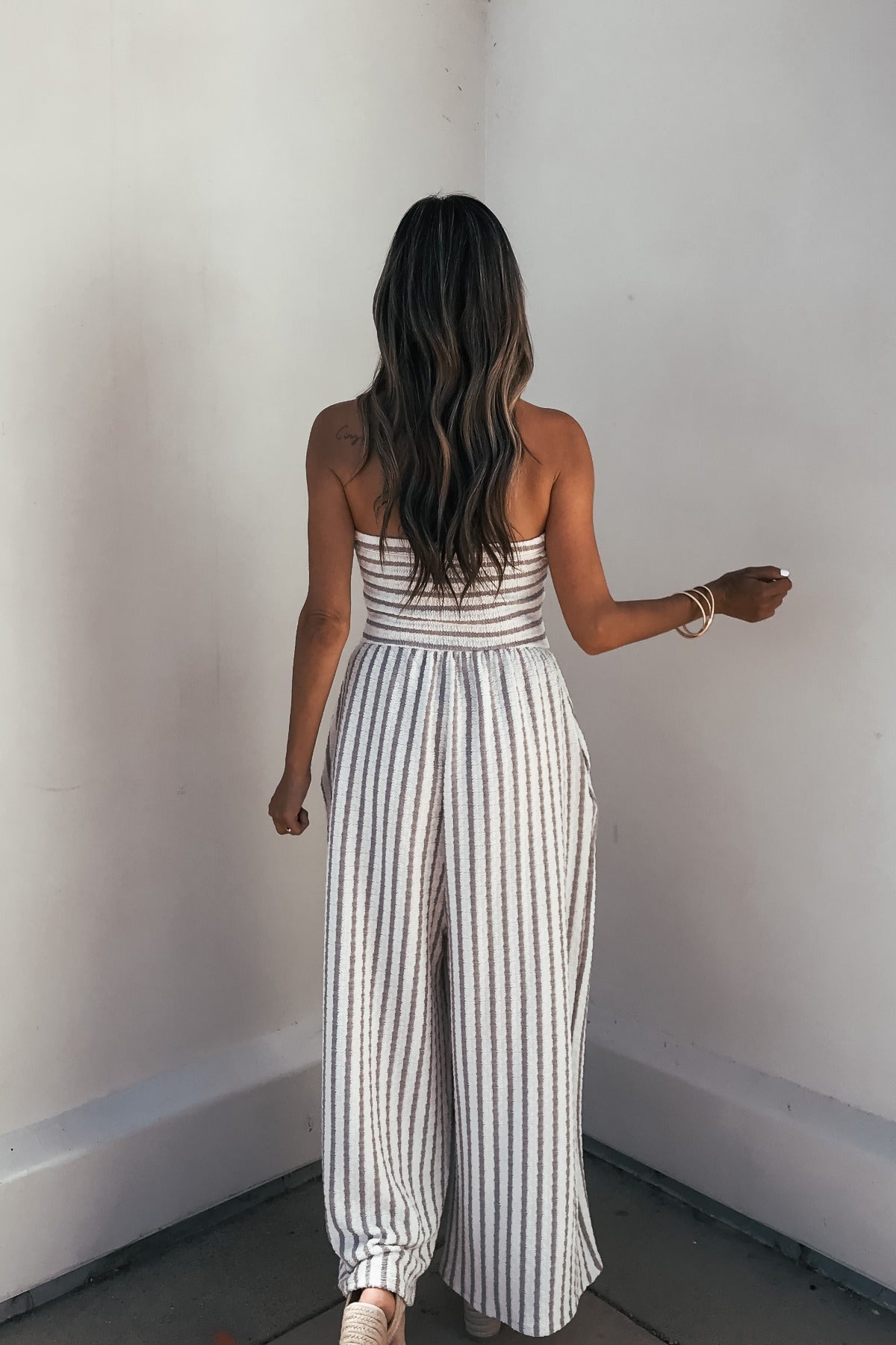 Woman with long wavy hair wears the Coastal Striped Wide Leg Jumpsuit - Taupe, facing a blank white wall.