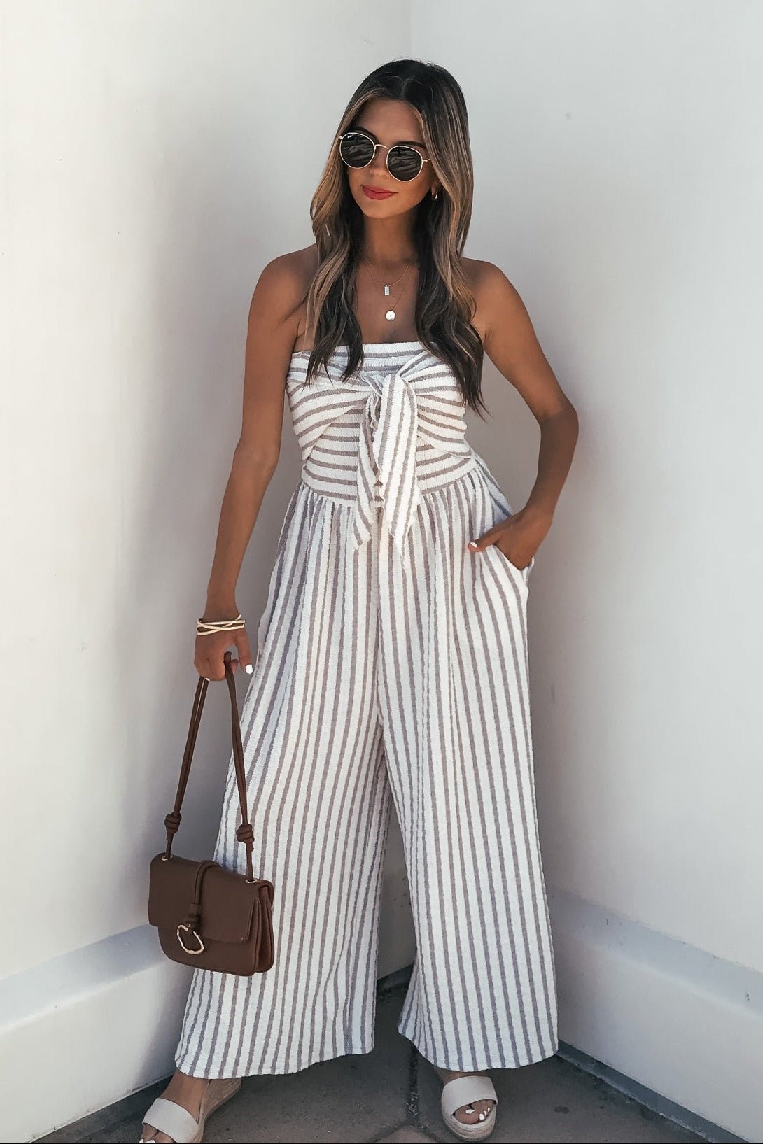 Woman in a Coastal Striped Wide Leg Jumpsuit - Taupe poses with a brown handbag, white sandals, and sunglasses by a white wall.