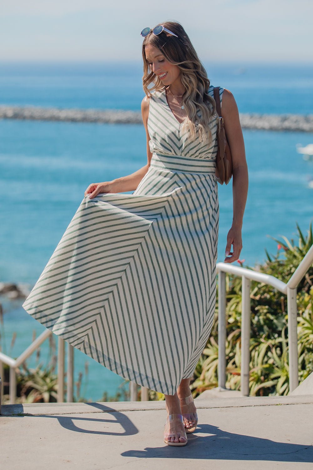 Woman wearing the Coastline Striped Midi Dress - Green stands on seaside stairs, smiling and holding her dress on a sunny day.