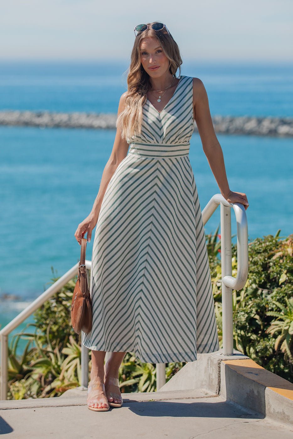 Woman in a Coastline Striped Midi Dress - Green stands by the ocean with a brown handbag and sunglasses.