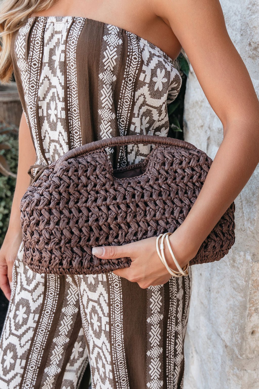 Woman in a patterned brown and white strapless jumpsuit holds the Cocoa Cabana Woven Clutch and wears silver bangles.