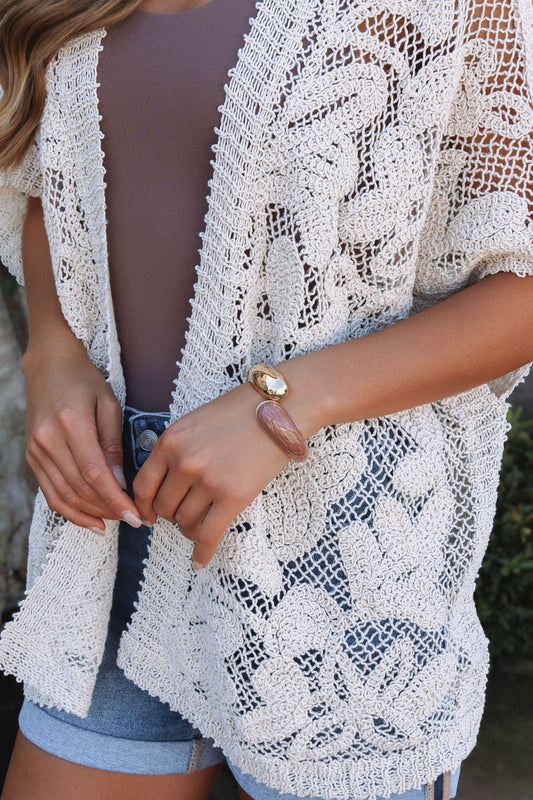 Woman in a beige crochet cardigan, mauve top, denim shorts, and the Cocoa Sol Hinge Bracelet with gold and pink stones.