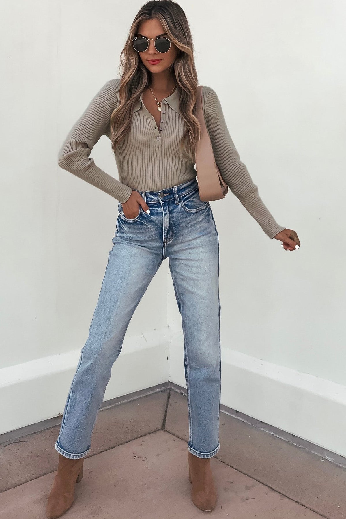 Woman in sunglasses, blue jeans, tan boots, and a Coffee Ribbed V-Neck Button Up Top poses stylishly by a white wall.