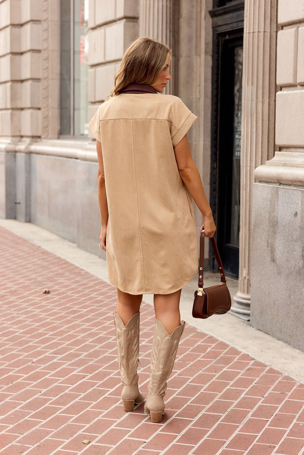 Woman in a tan Contrast Button Down Mini Dress stands on a brick sidewalk with cowboy boots, holding a brown purse, facing away.