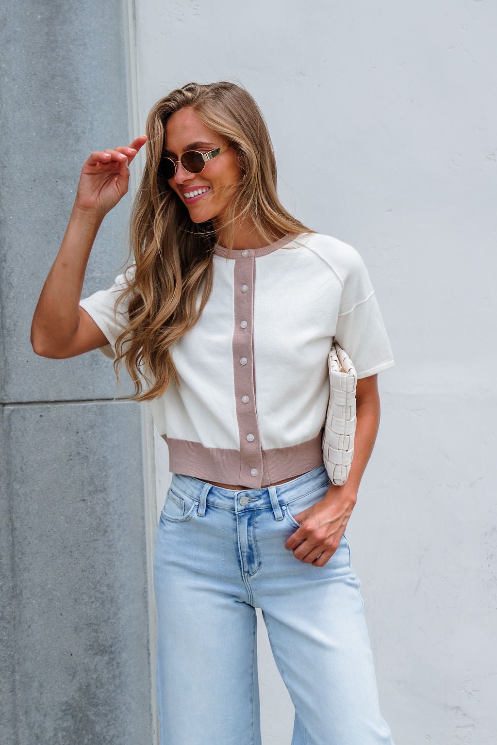 Woman in sunglasses smiles in a relaxed fit, cream Contrast Button Front Sweater and light jeans, holding a white clutch.
