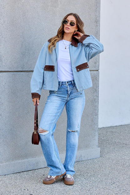Woman wears the Contrast Trim Jacket in dusty blue, white tee, ripped jeans, and sunglasses by a gray wall holding a brown purse.