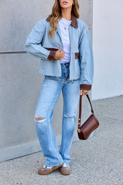 A person in a Contrast Trim Jacket - Dusty Blue (pre order), white shirt, ripped jeans, and brown shoes holds a handbag by a gray wall.