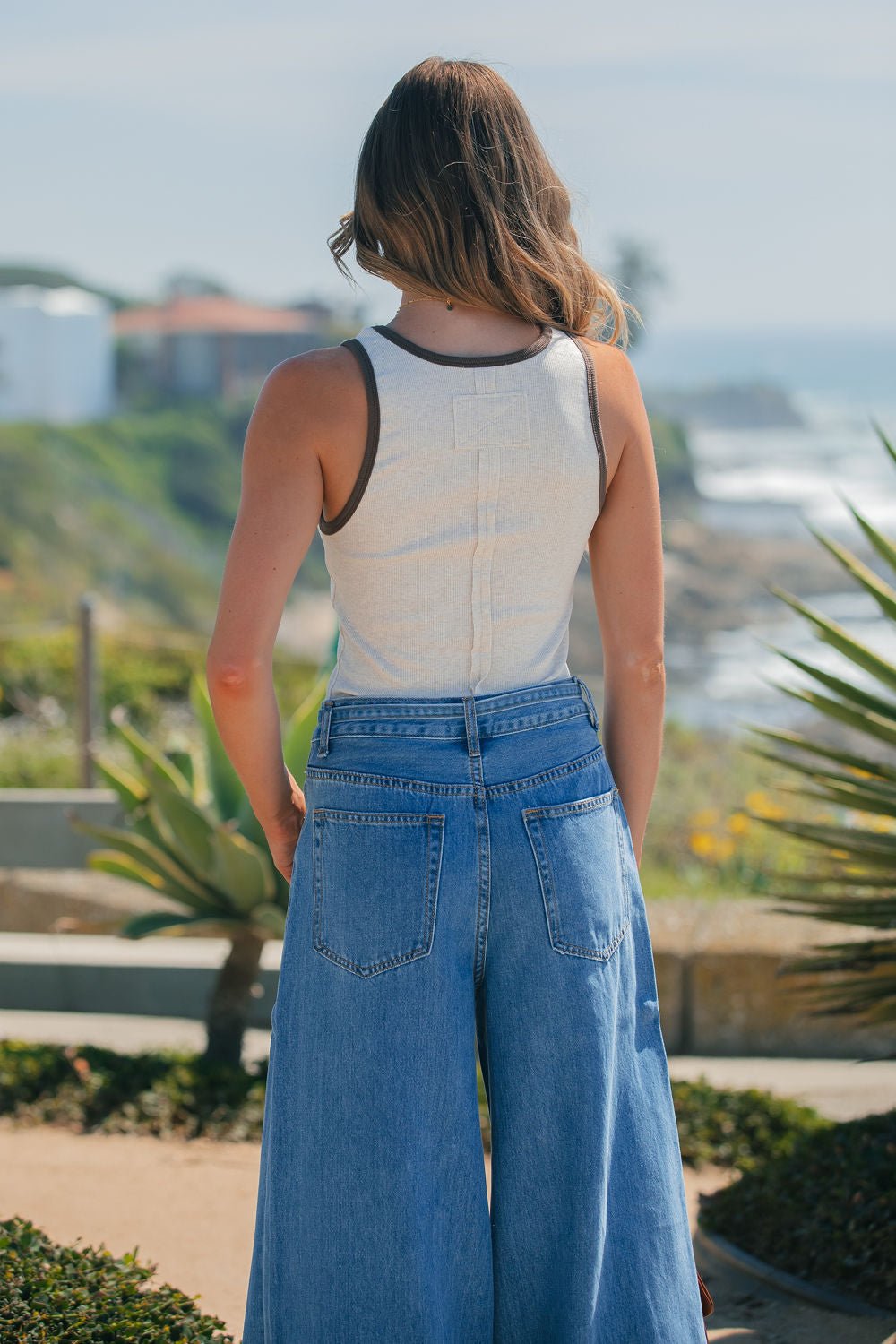 A woman with wavy hair faces away outdoors by the coast, wearing an Ivory Contrast Trim Tank Top and wide-leg jeans.