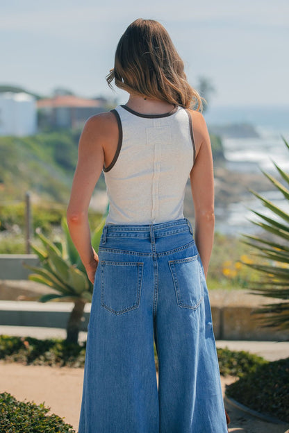 A woman with wavy hair faces away outdoors by the coast, wearing an Ivory Contrast Trim Tank Top and wide-leg jeans.