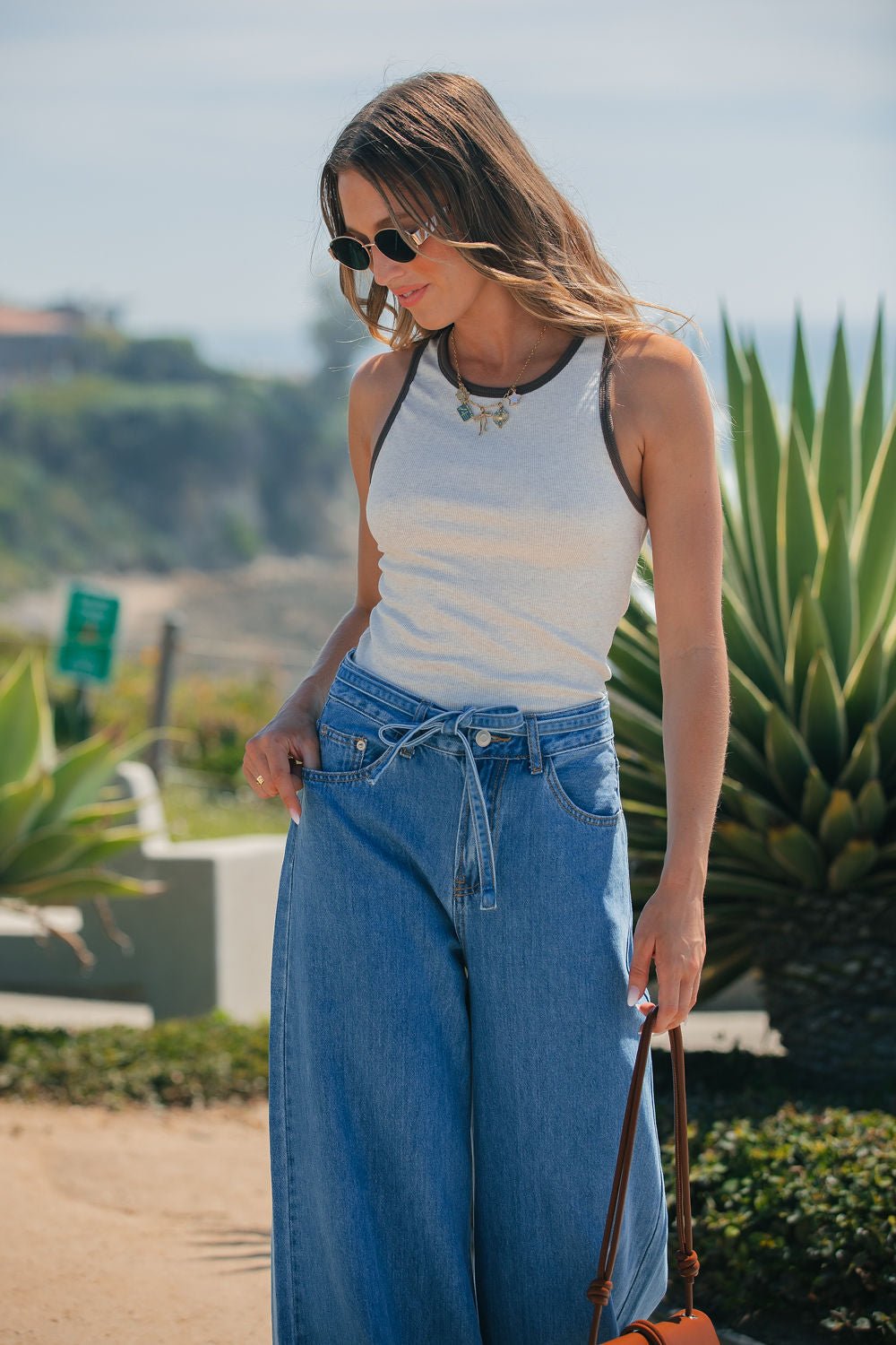 Woman in sunglasses wears an Ivory Contrast Trim Tank Top with wide-leg jeans, holding a brown bag outdoors by greenery.