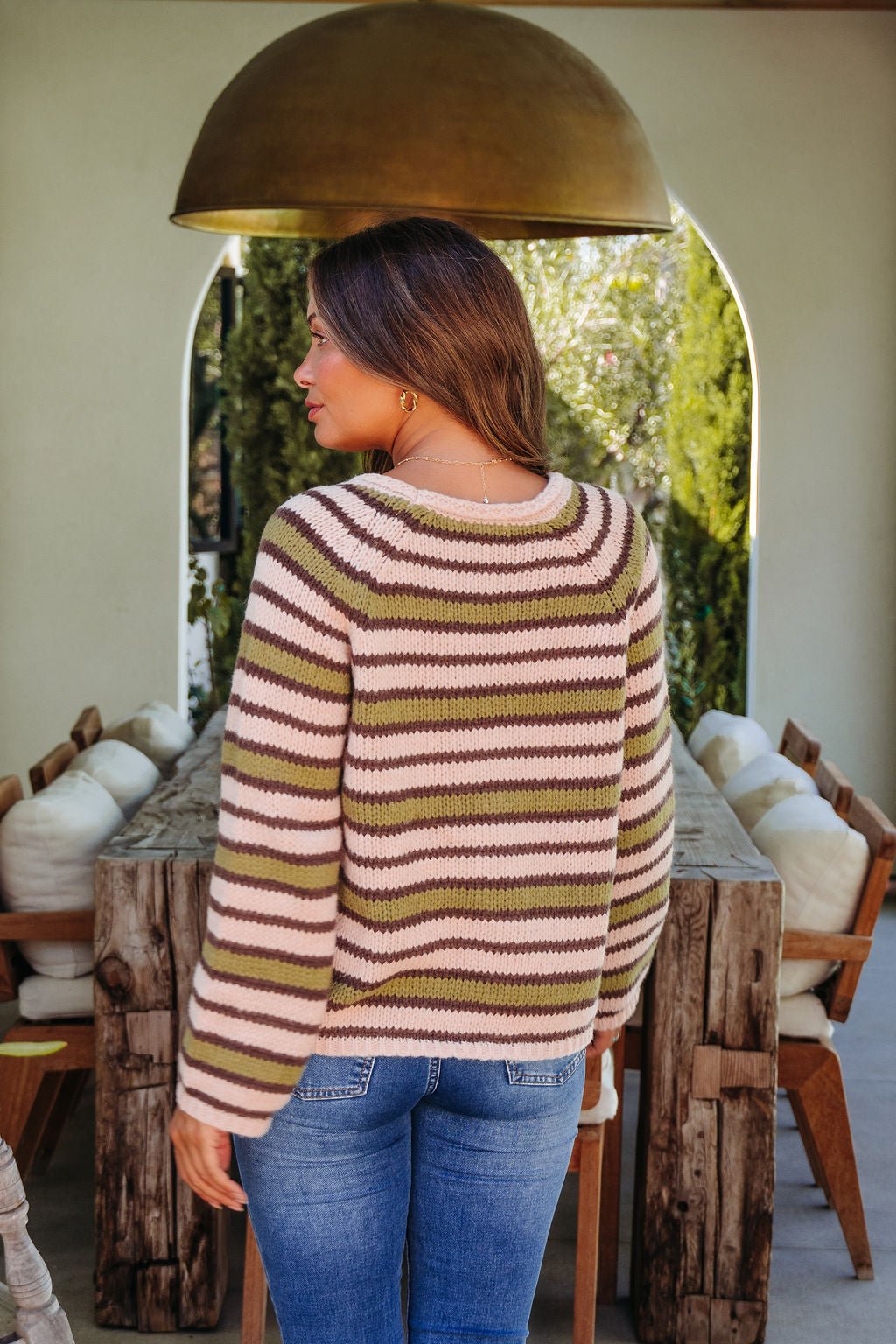 Woman with brown hair in the Corinne Multi Olive Striped Sweater stands by a rustic outdoor wooden table and chairs, facing away.