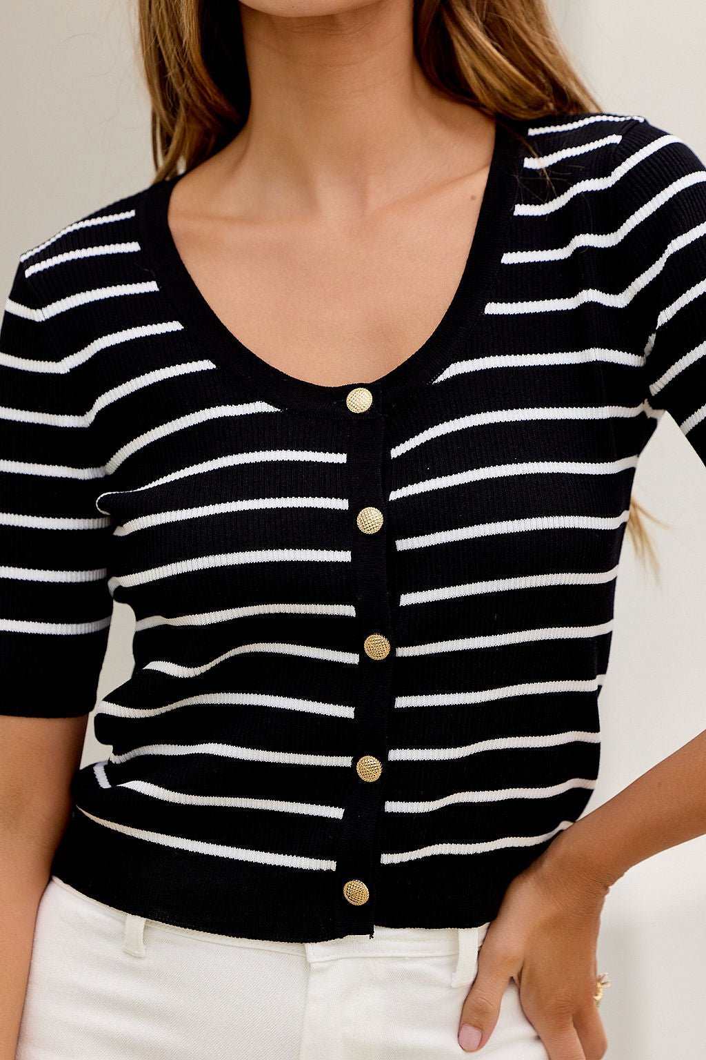 Woman wears the Corner Office Striped Top - Black with gold buttons, scoop neck, and white pants, hand on hip.