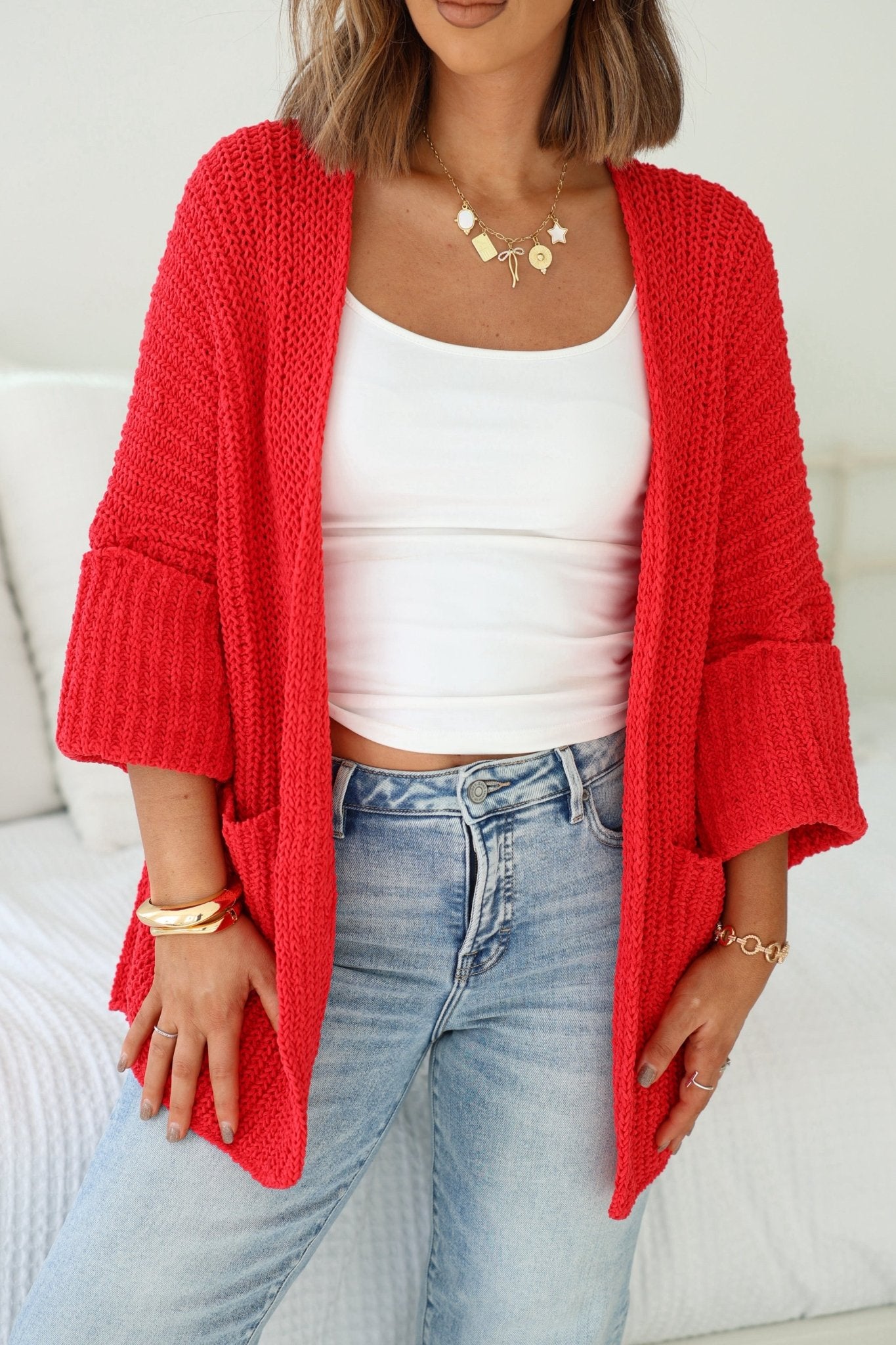 A woman wears a Cozy Apple Red Open Front Cardigan with a white tank top, blue jeans, and gold jewelry indoors.