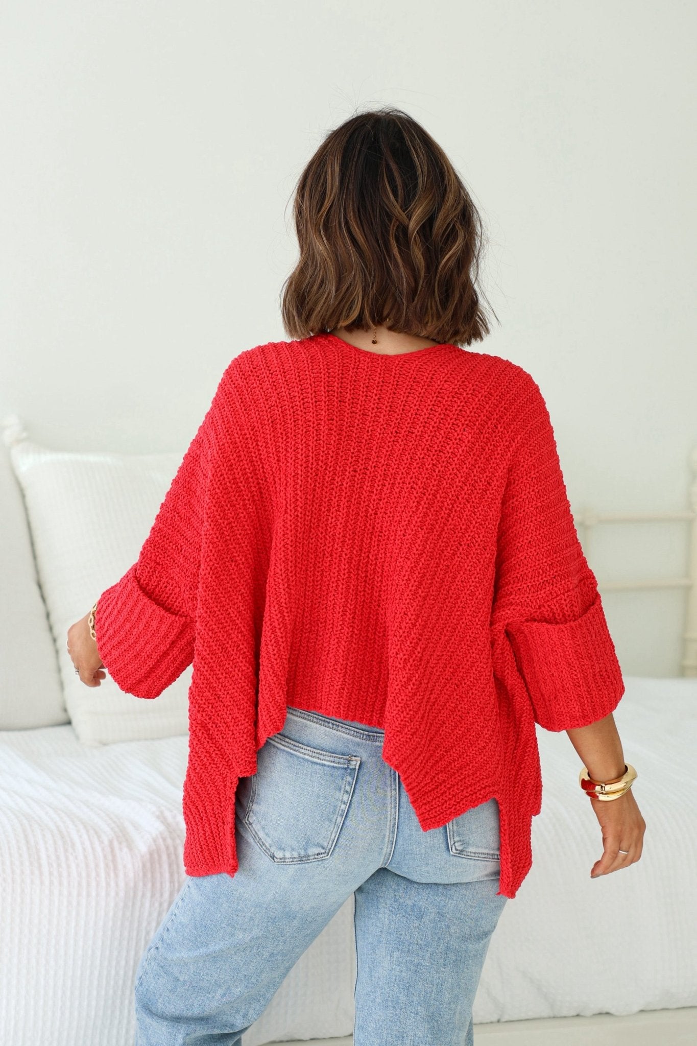 A person with wavy brown hair models the Cozy Apple Red Open Front Cardigan and light blue jeans, facing away in a bright room.