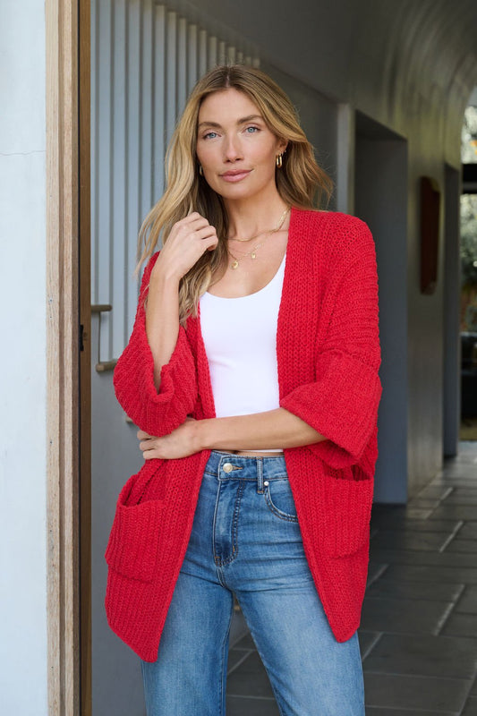Woman with long blonde hair wears the Cozy Apple Red Open Front Cardigan over a white top and blue jeans by an open doorway.