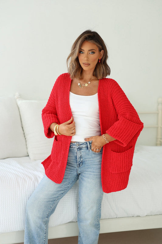 Woman with shoulder-length hair in a Cozy Apple Red Open Front Cardigan, white top, and jeans poses indoors in a bright bedroom.
