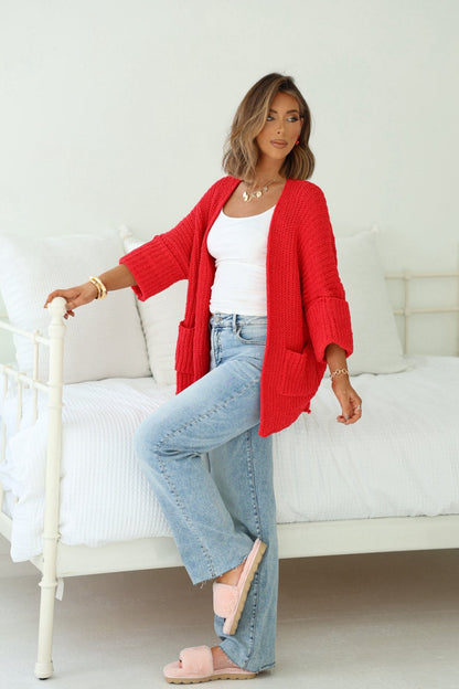 Woman in Cozy Apple Red Open Front Cardigan, white tank, and jeans poses by a bed, lifting one leg in pink slippers.