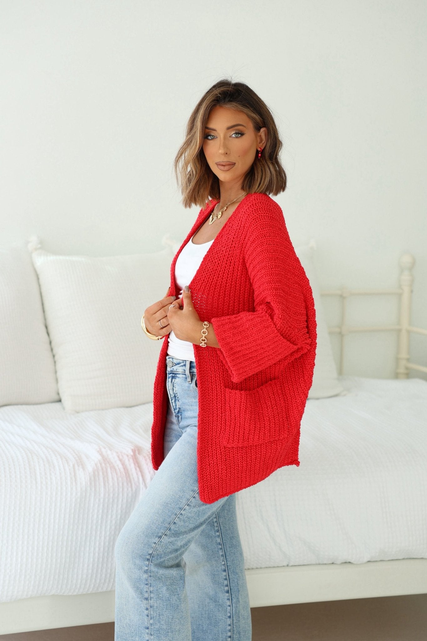 A woman in a Cozy Apple Red Open Front Cardigan and blue jeans stands by a white bed in a bright, inviting room.