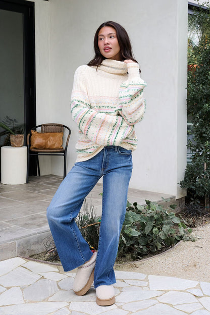 A woman poses outdoors in the Cozy Cream Turtleneck Striped Sweater and blue jeans, standing on a stone path.
