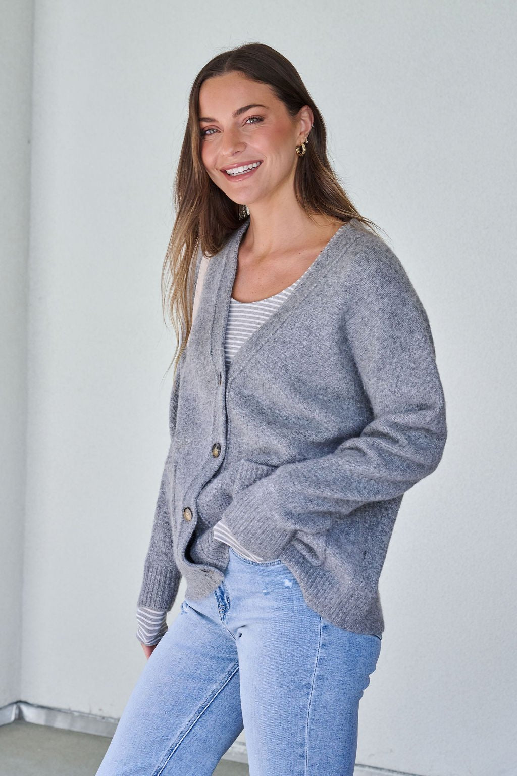 Smiling woman with long brown hair wears the Cozy Grey Boyfriend Cardigan, striped shirt, and light blue jeans against a white wall.