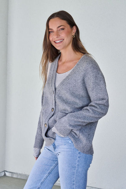 Smiling woman with long brown hair wears the Cozy Grey Boyfriend Cardigan, striped shirt, and light blue jeans against a white wall.