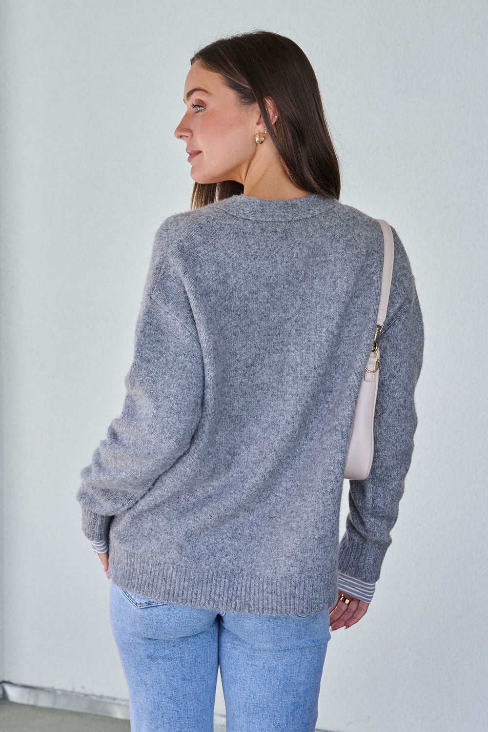 A woman with long brown hair wears the Cozy Grey Boyfriend Cardigan - FINAL SALE, light blue jeans, and holds a white shoulder bag.