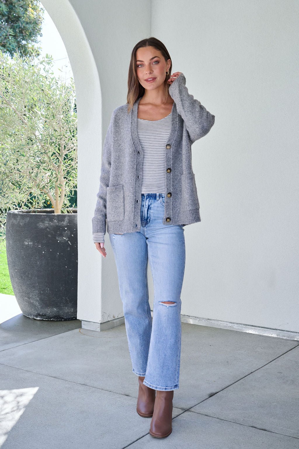 Woman outside in a Cozy Grey Boyfriend Cardigan, striped top, light blue jeans, and brown ankle boots, hand in her hair.