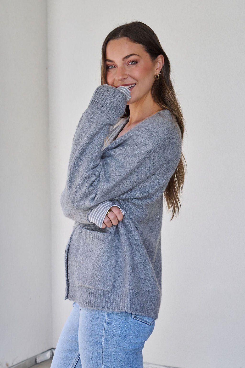 A woman smiles, posing in a Cozy Grey Boyfriend Cardigan and light blue jeans against a plain background.