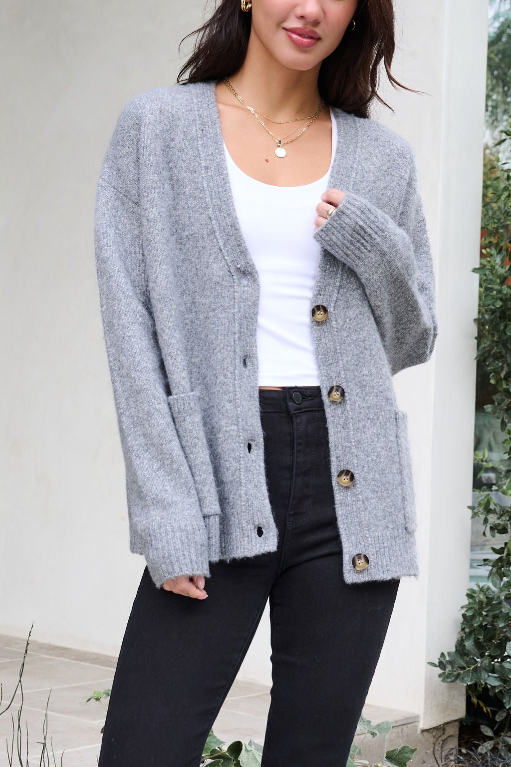 Woman in a Cozy Grey Boyfriend Cardigan over a white top and black pants, standing outdoors near greenery.