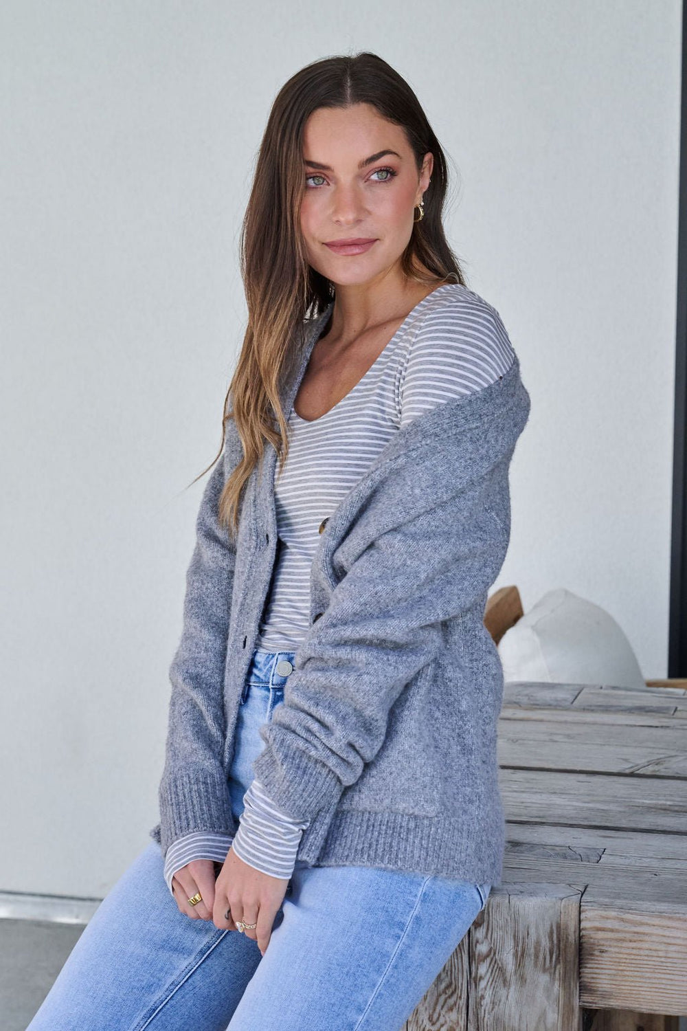 Woman with long brown hair wears the Cozy Grey Boyfriend Cardigan - FINAL SALE, sitting on a wooden bench and smiling softly.