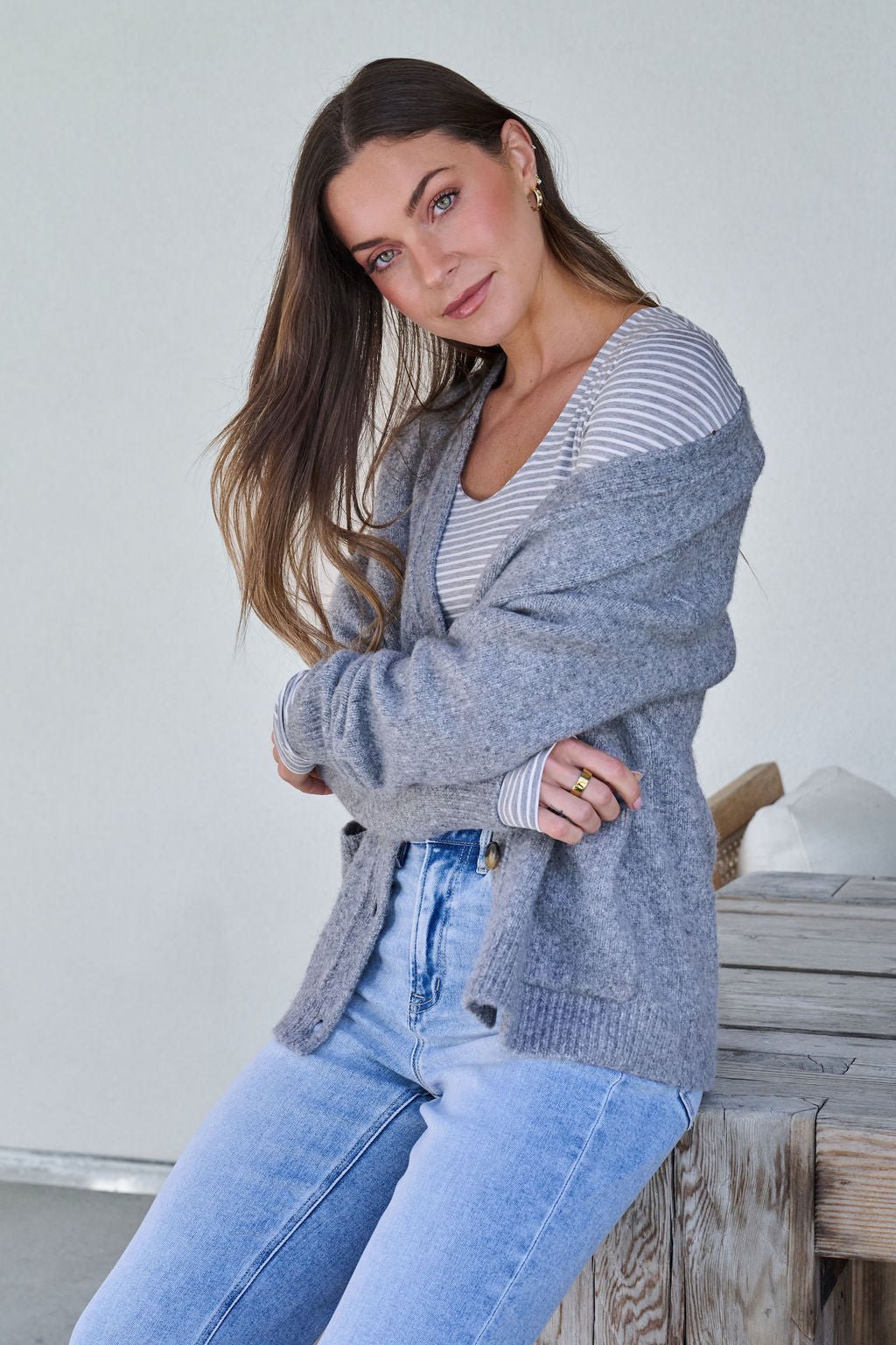 A woman with long brown hair wears the Cozy Grey Boyfriend Cardigan and sits on a wooden bench, looking at the camera.