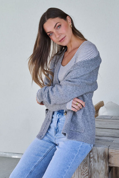 A woman with long brown hair wears the Cozy Grey Boyfriend Cardigan and sits on a wooden bench, looking at the camera.