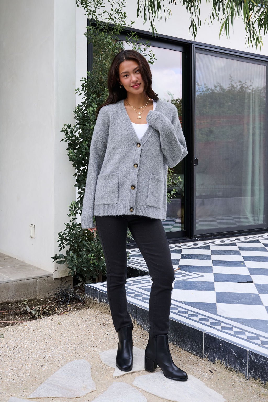 Woman outside on a checkered patio, wearing the Cozy Grey Boyfriend Cardigan, white top, black pants, and black boots.